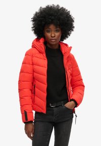 Rode pufferjack met capuchon, doorgestikt ontwerp en zijrits zakken. Draag het over een zwarte trui en gestyled met donkere spijkerbroeken.
