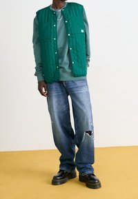 Chaleco acolchado verde con broches plateados sobre un sudadera gris, combinado con jeans azules desgastados y botas negras gruesas. Capas texturizadas visibles.