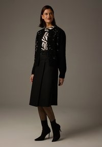 Schwarze Strickjacke mit strukturierten Akzenten über einem weißen Hemd mit einem schwarzen abstrakten Muster. Ein schwarzer Midi-Rock und Stiefeletten runden den Look ab.