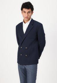 Marineblaues, doppelt-knöpfbares Blazer mit zwei vorderen Taschen und aufwendigen goldenen Knöpfen. Begleitet von einem weißen Hemd und hellblauen gemusterten Hosen.