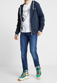 Sudadera con cremallera azul marino y cordones blancos, combinada con una camiseta blanca con logo negro, jeans azules y zapatillas gruesas multicolores.