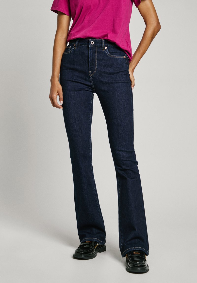 Jeans acampanados de un azul oscuro con cintura alta, textura de mezclilla, costuras naranjas y cierre frontal con botón, combinados con una camiseta rosa.