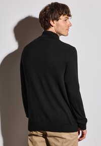 Pull noir à col montant, avec des poignets côtelés et une coupe décontractée. Texture lisse, porté avec un pantalon beige, montrant uniquement la vue arrière.