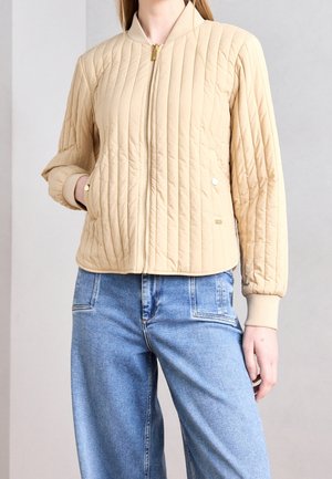 Femme portant une veste matelassée beige zippée avec des poignets côtelés et un jean bleu clair taille haute, debout sur un fond uni.