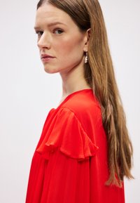 Vestido de tela roja con mangas en capas y volantes, presentando una silueta fluida. La modelo tiene el cabello largo y lleva pendientes parecidos a perlas.