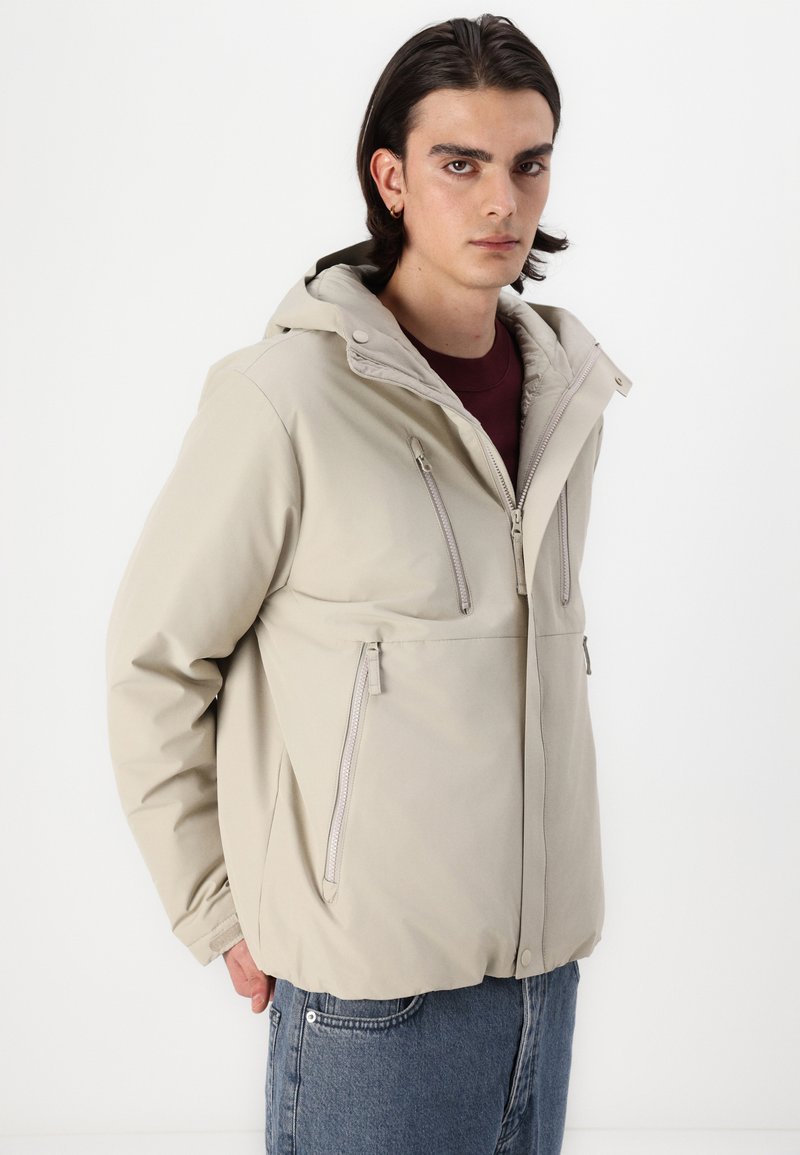 Chaqueta impermeable beige con capucha, dos bolsillos en el pecho con cremallera y bolsillos laterales. Presenta una textura suave y un ajuste relajado.