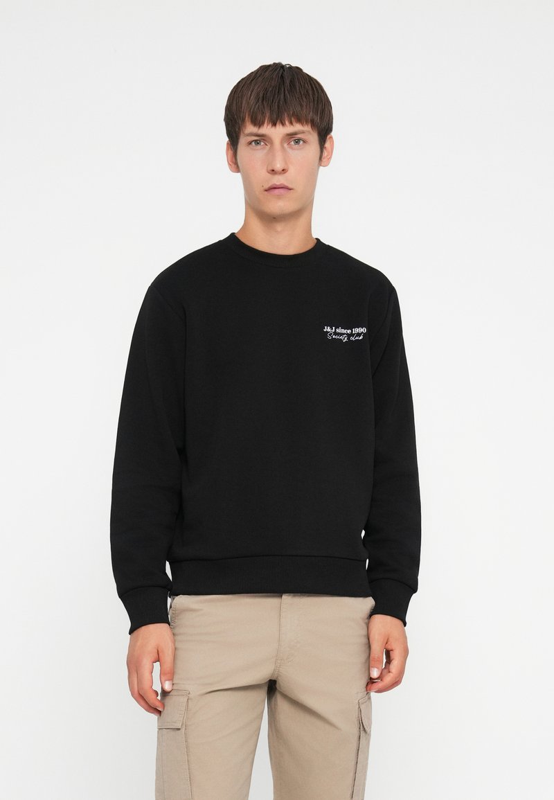 Sweat-shirt noir à manches longues, avec des poignets et un ourlet côtelés, arborant une broderie blanche sur la poitrine avec l'inscription "J&J depuis 1990."