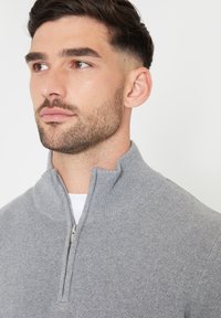 Grijze zip-up sweater met een hoge kraag en geribbelde textuur. Heeft een metalen rits bij de hals en een gladde, lichte stof.