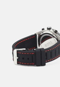 Correa de reloj de silicona negra con costuras rojas, hebilla plateada y caja de reloj metálica visible al final. Presenta diales multifunción.