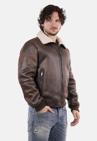 Giubbotto bomber in pelle marrone con colletto in shearling, zip frontale e due tasche. I polsini e il bordo a coste aggiungono texture e forma.