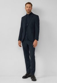 Maßgeschneiderter dunkelblauer Anzug mit einem Reißverschlusskragen, taillierten Hosen und glatter Textur. Getragen mit schwarzen Schuhen vor einem hellgrauen Hintergrund.