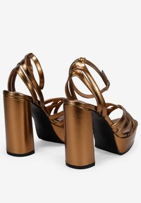 Sandales à talons hauts métalliques en bronze avec de larges talons carrés, des lanières croisées et des brides autour de la cheville. Fini lisse, design ouvert aux orteils.