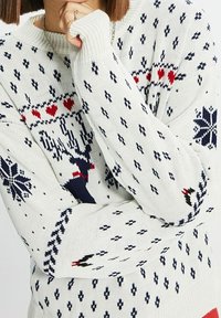 Vit stickad tröja med olika mönster i marinblått och rött, inklusive snöflingor, hjärtan och renar, med ribbade ärmslut och halsringning.