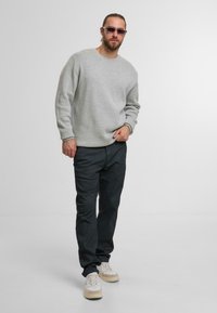 Felpa grigia con collo rotondo, vestibilità ampia; abbinata a pantaloni chinos scuri e sneakers chiare. Gli accessori includono occhiali da sole sottili.