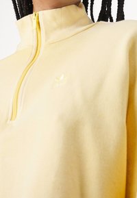 Keltainen vetoketjullinen collegepaita korkealla kauluksella, valmistettu pehmeästä kankaasta, jossa on vastaava vetoketju ja vasemmassa rinnassa kohokuvioitu logo.