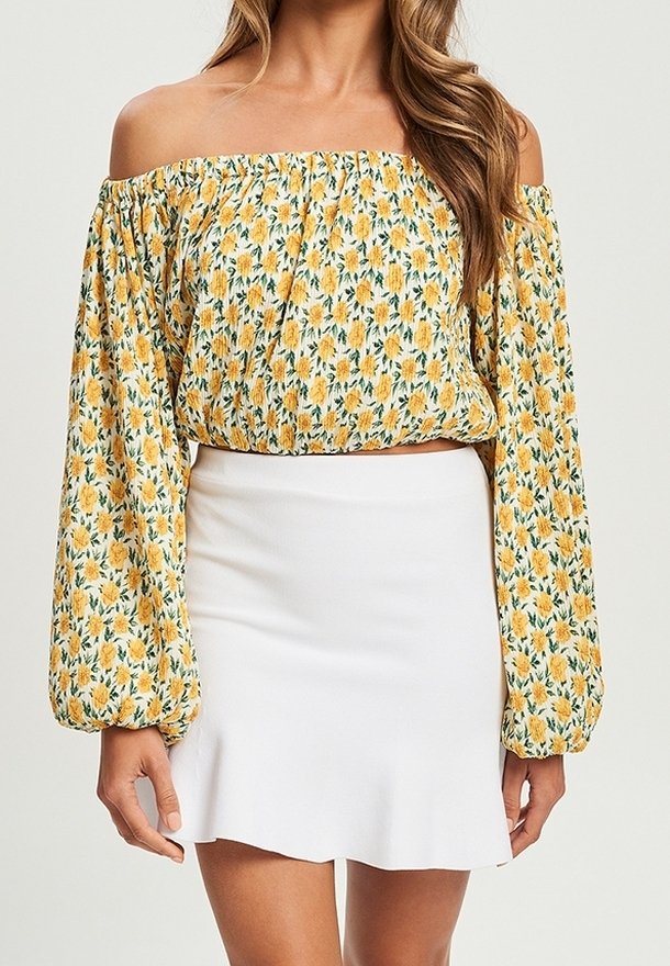 Femme portant un chemisier jaune fleuri à épaules dénudées avec manches longues et une jupe blanche taille haute.