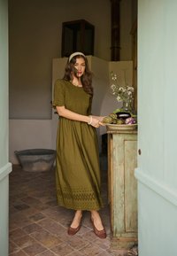 Ženska v olivno zeleni obleki in rjavih čevljih stoji blizu rustikalne omare s krožnikom zelenjave in cvetja v slabo osvetljeni sobi.