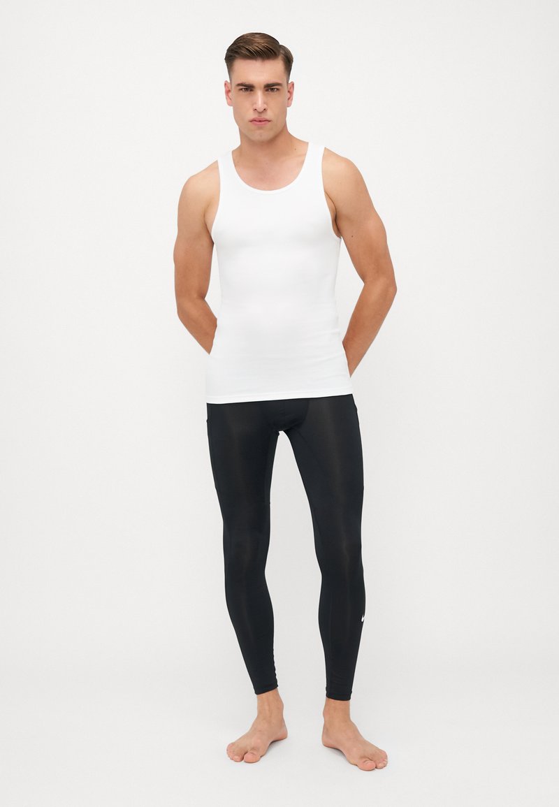 Witte mouwloze tanktop gedragen met zwarte legging. De tank is glad en aansluitend; de leggings zijn rekbaar en sleek, met een klein logo.