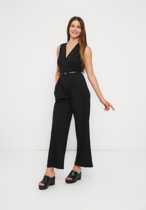 Mosolygó nő hosszú barna hajjal, fekete ujjatlan overált övvel és fekete platform szandállal, fehér háttér előtt áll.