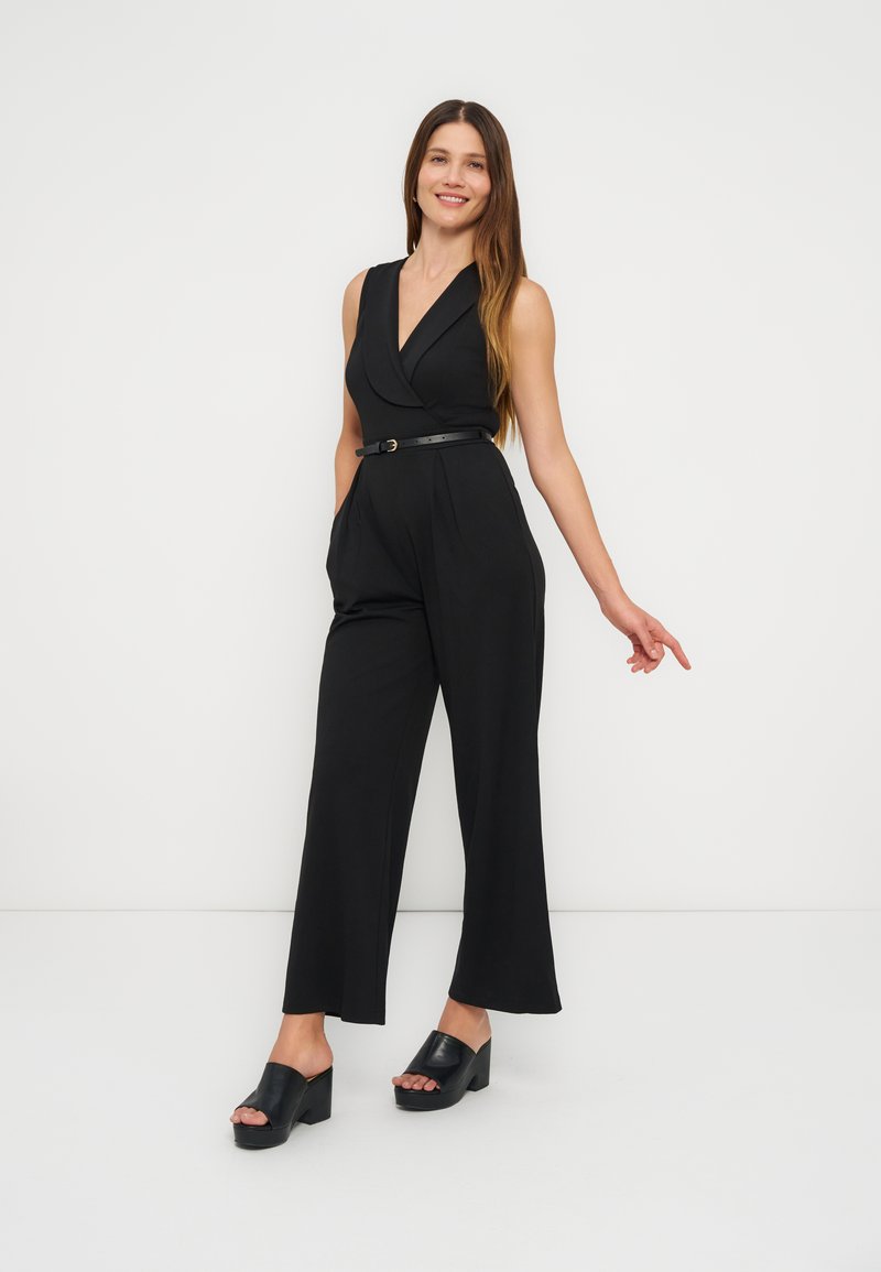 Femme souriante aux longs cheveux bruns portant une combinaison noire sans manches avec ceinture et des sandales plateformes noires, debout devant un fond blanc.