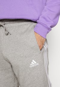 Grå bomullssvettshorts med vitt Adidas-logotyp, sidofickor och elastiskt midjeband. Matchas med en lila sweatshirtärm.