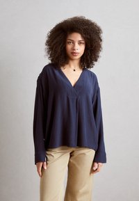 Blusa lunga navy a maniche lunghe con scollatura a V, vestibilità rilassata e tessuto testurizzato. Abbinata a pantaloni beige chiaro a gamba dritta.