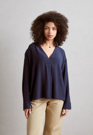 Blusa lunga navy a maniche lunghe con scollatura a V, vestibilità rilassata e tessuto testurizzato. Abbinata a pantaloni beige chiaro a gamba dritta.