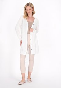 Weißer langer Cardigan mit Knopfverschluss, aus leichtem Stoff, getragen über einem beigen Top, kombiniert mit beigen cropped Hosen und hellem Schuhwerk.