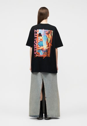 Kobieta o długich włosach nosi czarny T-shirt z kolorową, vintage'ową sceną plażową, w połączeniu z jasną spódnicą jeansową z tylnym rozcięciem i czarnymi butami.