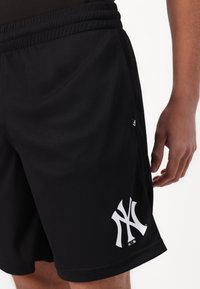 Pantalones cortos deportivos negros con cintura elástica, con un gran logo blanco a la izquierda y una pequeña etiqueta en el costado. Textura suave.