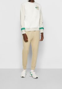 Sweatshirt i vitt med grönrandiga muddar och en beige logotyp, tillsammans med beige sweatpants och vita sneakers med gröna detaljer.