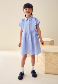 lichtblauw geruit jurkje met korte ruches mouwen, knoopsluiting aan de voorkant en een geplooide taille. G gedragen met zwarte schoenen en witte sokken.