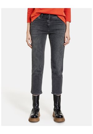 Femme portant un jean noir délavé à taille moyenne, court, avec des bottes en cuir noires et un haut à manches longues orange, vue de la taille aux pieds.