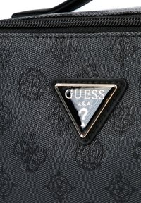 Bolso de material negro texturizado con un logo triangular de herrajes dorados que incluye "GUESS" y un signo de interrogación decorativo. Cierre con cremallera en la parte superior.