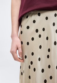 Main d'une personne reposant à côté d'une jupe beige à pois noirs, portant un haut bordeaux.