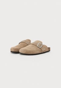 Brown suede clogs with a rounded toe, featuring a wide strap with silver hardware and circular cutouts, resting on a textured black sole.