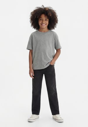 Smilende barn med krøllete hår iført grå t-skjorte, svarte jeans, hvite joggesko, står med en hånd i lommen på hvit bakgrunn.