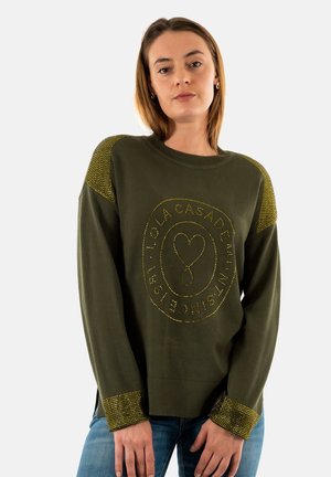 Sudadera verde oliva con cuello y puños acanalados, que presenta un logo de tachuelas amarillas en la parte delantera. Aberturas en los costados del dobladillo.