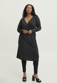 Schwarzes Wickelkleid mit tiefem V-Ausschnitt, langen Ärmeln und strukturiertem Stoff. Kombiniert mit schwarzen Leggings und Plattformsandalen mit glatter Oberfläche.