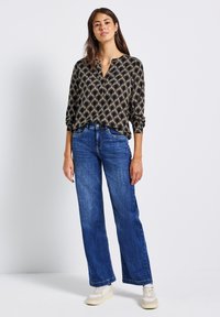 Donna che indossa una blusa a maniche lunghe nera e beige con un motivo, jeans a gamba larga blu e sneakers bianche, in piedi contro uno sfondo bianco semplice.