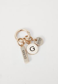 Gullfarget nøkkelring med tre anheng: en rektangulær plate med "GUESS," en sirkulær plate med et "G," og et lite rundt merke merket "EST. 1981."