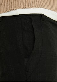 Detailní záběr na kapsu a pas černých kostkovaných kalhot s béžovým úpletem zasunutým dovnitř, který zdůrazňuje texturu látky a detaily šití.