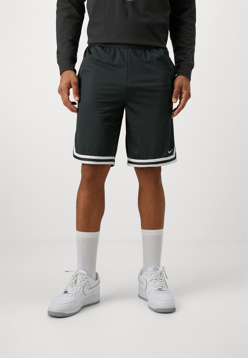 Homme portant un short de basket noir avec des rayures blanches, des chaussettes blanches montantes et des baskets blanches Nike, debout devant un fond uni.