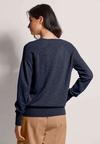Frau mit dunklem Haar in einem navyblauen Langarmpullover und beigen Hosen steht mit dem Rücken zur Kamera, eine Hand auf der Hüfte, vor einem schlichten Hintergrund.
