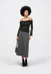 Femeie cu coafură afro, purtând un top negru cu umerii goi, cu mâneci transparente, fustă gri midi, șosete negre, încălțăminte și o poșetă închisă la culoare.