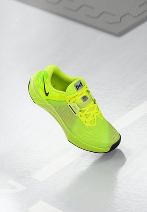 Chaussure de course jaune-vert néon vif avec semelle et lacets noirs, posée sur un sol texturé gris clair.