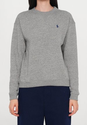 Personne portant un pull gris uni à col rond avec un petit logo bleu brodé sur le côté gauche de la poitrine et un pantalon bleu foncé.