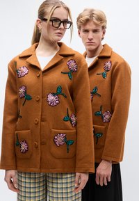 Καφέ μάλλινα σακάκια με κεντήματα από λουλουδάτα παγιέτες, με χαραγμένους γιακάδες και εμπρόσθιες τσέπες, που φοριούνται από δύο μοντέλα σε ουδέτερο φόντο.