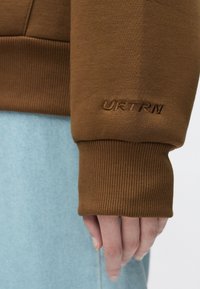 Ruda džemperis su siūtais „URTRN“ užrašu ant riboto rankogalių, derinamas su šviesiai mėlynais džinsais. Artimas rankovės ir rankos detalės vaizdas.