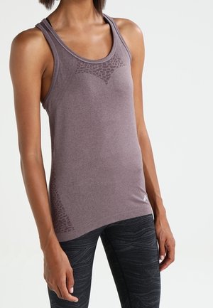 Femme portant un débardeur de sport violet sans manches et un legging noir à motifs, posant devant un fond clair uni.